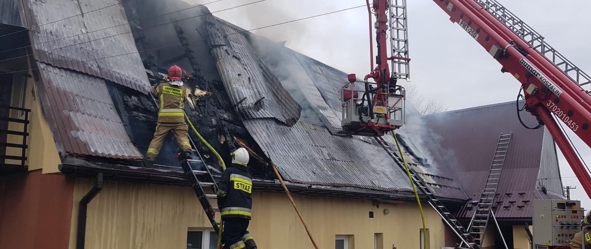 Zdjęcie przedstawia pracujących strażaków podczas gaszenia pożaru w miejscowości Budzów. Strażacy walczą z pożarem dachu.