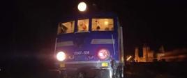 Zdjęcie przedstawia przód lokomotywy pociągu pasażerskiego, lokomotywa koloru niebieskiego z dwoma okrągłymi światłami na przodzie. W tle krajobraz nocny. 