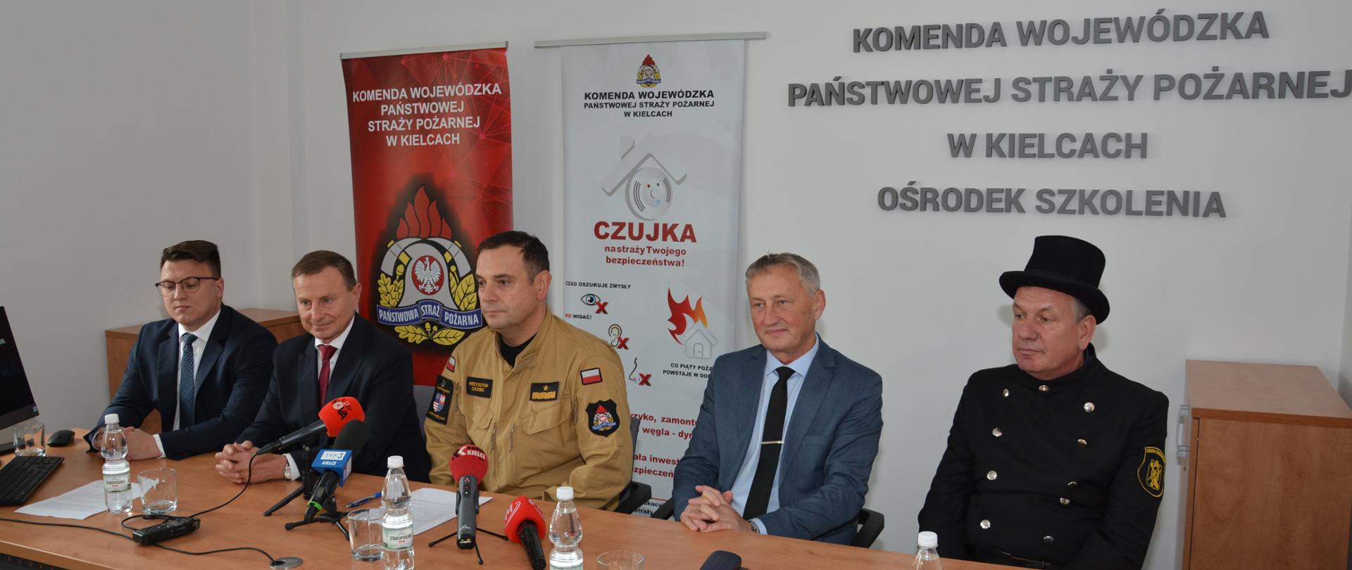 Na zdjęciu widzimy 5 mężczyzn siedzących podczas konferencji prasowej. W środku mężczyzna w mundurze piaskowym PSP, a pierwszy od prawej siedzi kominiarz. Pozostali trzej mężczyźni w garniturach. 