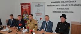Na zdjęciu widzimy 5 mężczyzn siedzących podczas konferencji prasowej. W środku mężczyzna w mundurze piaskowym PSP, a pierwszy od prawej siedzi kominiarz. Pozostali trzej mężczyźni w garniturach. 