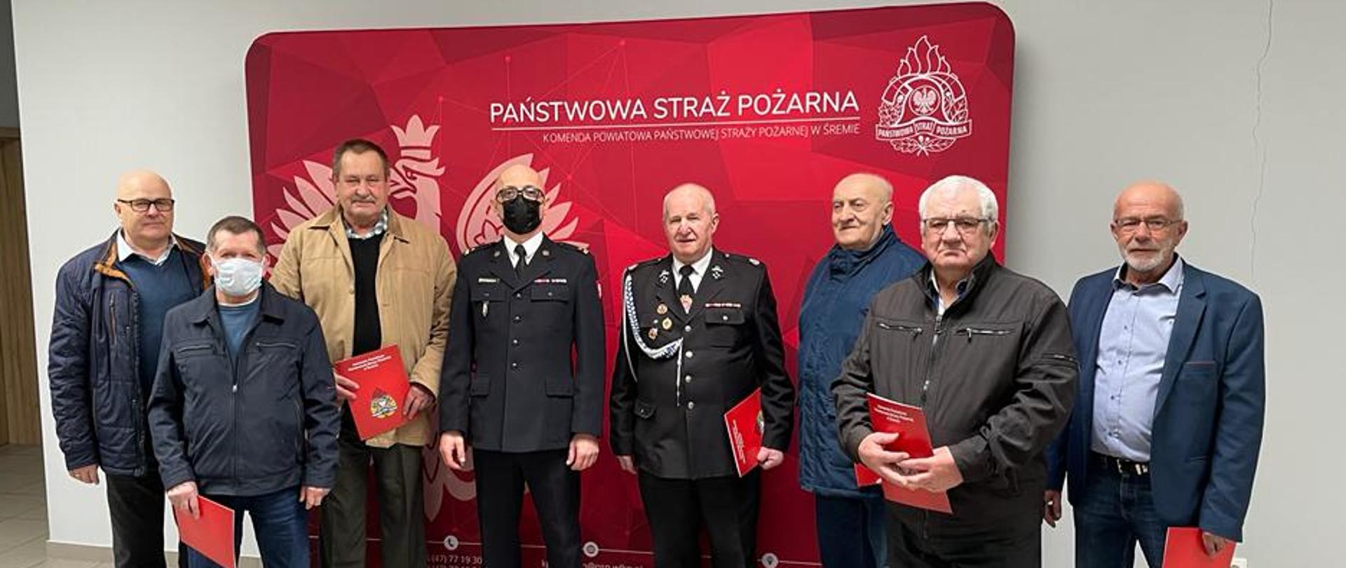 Na zdjęciu druhowie OSP z wręczonymi decyzjami z komendantem Powiatowym PSP na tle ścianki z godłem i logiem PSP oraz napisem KPPSP w Śremie 