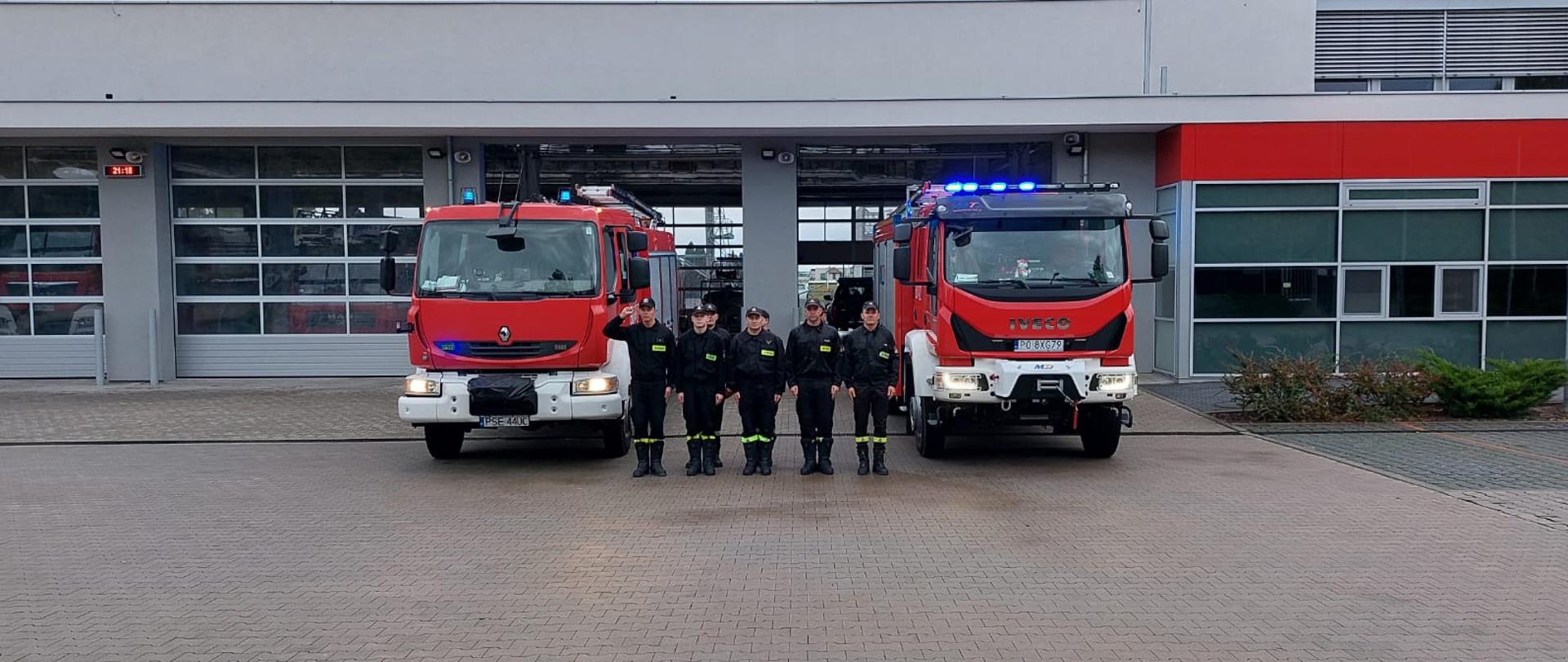 Na zdjęciu strażacy udający hołd zmarłemu koledze przed bramami garażowymi stoją dwa samochody ratowniczo-gaśnicze z włączonymi sygnałami świetlnymi przed nimi w dwuszeregu stoją strażacy 
