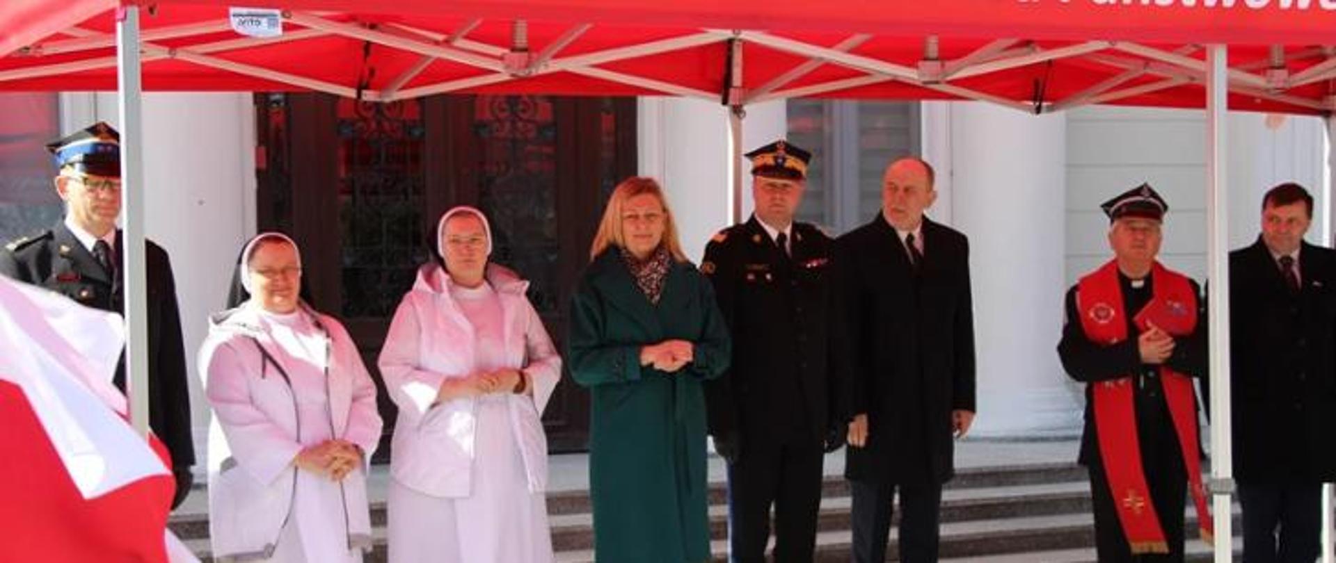 grupa ludzi w tym strażacy, ksiadz i siostry zakonne stoją pod namiotem