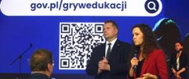 Minister Czarnek i kobieta w czerwonej marynarce stoją przed ekranem, na którym na niebieskim tle jest kod QR i napis gov.pl/grywedukacji.