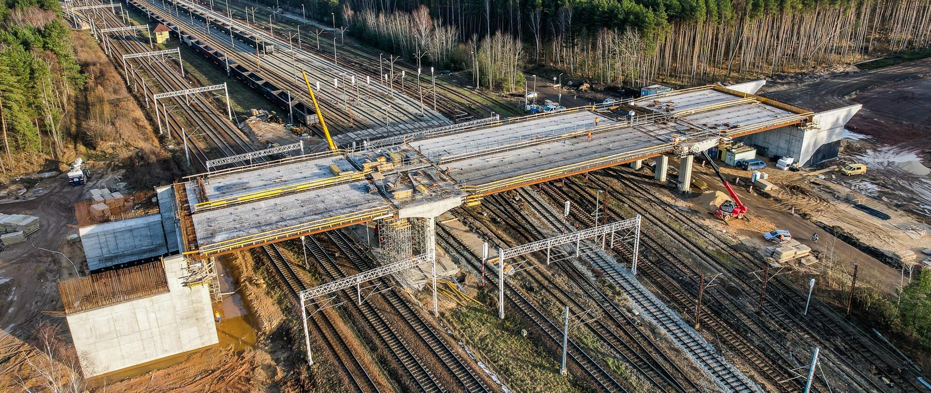 Wiadukt drogowy w budowie. Widoczne szalunki pomostu wiaduktu, oraz pracownicy i maszyny budowlane wspomagające budowę obiektu.