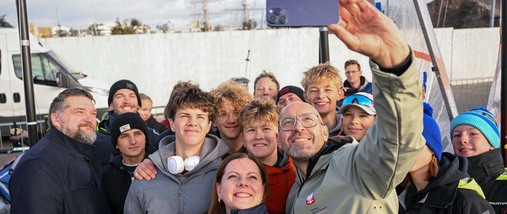 Minister sportu i turystyki robi sobie selfie z prezydent Gdyni Aleksandrą Kosiorek i grupą młodych osób. W tle widać maszty żaglówek.