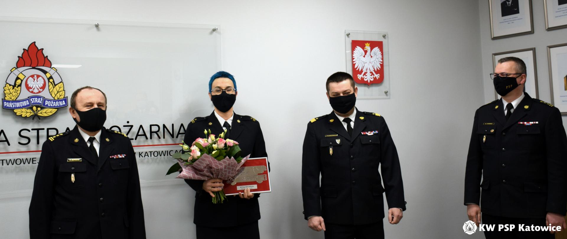 Zdjęcie przedstawia Śląskiego Komendanta Wojewódzkiego PSP nadbryg. Jacka Kleszczewskiego, jego Zastępców - st. bryg. Bogdana Jędrochę i st. bryg. Mirosława Synowca, stojących z mł. bryg. Martą Różycką, która w ręce trzyma bukiet kwiatów i pamiątkową tabliczkę. Zdjęcie wykonano w sali sztabowej Komendy Wojewódzkiej PSP w Katowicach. Wszystkie osoby na zdjęciu są w mundurach wyjściowych i maseczkach ochronnych .