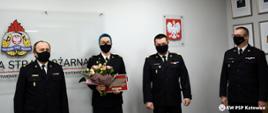 Zdjęcie przedstawia Śląskiego Komendanta Wojewódzkiego PSP nadbryg. Jacka Kleszczewskiego, jego Zastępców - st. bryg. Bogdana Jędrochę i st. bryg. Mirosława Synowca, stojących z mł. bryg. Martą Różycką, która w ręce trzyma bukiet kwiatów i pamiątkową tabliczkę. Zdjęcie wykonano w sali sztabowej Komendy Wojewódzkiej PSP w Katowicach. Wszystkie osoby na zdjęciu są w mundurach wyjściowych i maseczkach ochronnych.