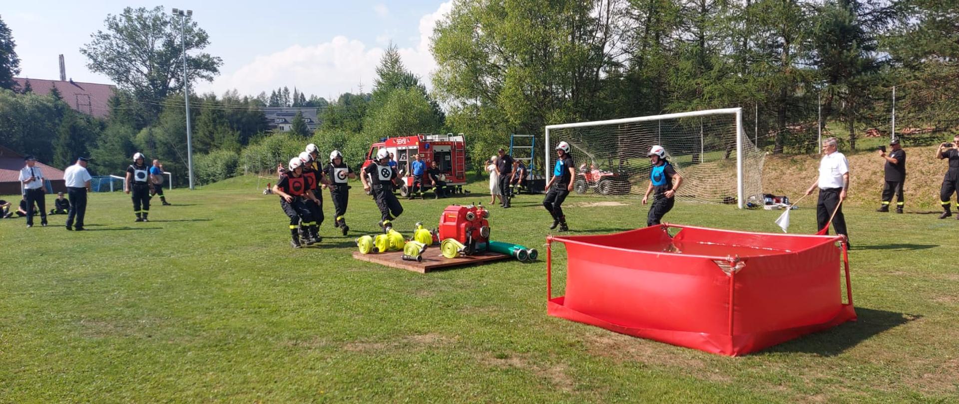 Strażacy biegnący w kierunku podestu na którym rozłożony jest sprzęt pożarniczy.