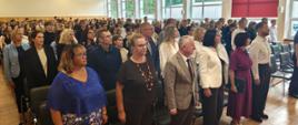 Uczniowie, rodzice i nauczyciele stojący na baczność w auli szkolnej podczas odśpiewania hymnu państwowego.