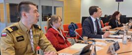 Zdjęcie przedstawia grupę osób siedzących przy stole konferencyjnym w sali spotkań. W pierwszym planie po lewej stronie widoczny jest mężczyzna w mundurze strażackim z naszywką „Państwowa Straż Pożarna” i imiennikiem „Mariusz Feltynowski”. Ma krótkie, ciemne włosy i skupiony wyraz twarzy. Obok niego siedzi kobieta w czerwonej marynarce i okularach, z włosami związanymi w kucyk. Dalej przy stole znajdują się inni uczestnicy spotkania, w tym mężczyzna w garniturze i kobieta z długimi rudymi włosami, pracujący przy laptopach i z dokumentami. Na stole znajdują się mikrofony, notatniki, butelki wody oraz tabliczki z nazwami krajów, w tym „Poland”. W tle widoczne są szklane kabiny tłumaczy oraz inni uczestnicy wydarzenia. Całość odbywa się w nowoczesnej sali konferencyjnej z drewnianymi panelami na ścianach.