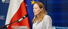 Minister funduszy i polityki regionalnej Katarzyna Pełczyńska-Nałęcz stoi przy mównicy i przemawia do mikrofonu. Po jej prawej stronie flagi Polski i UE