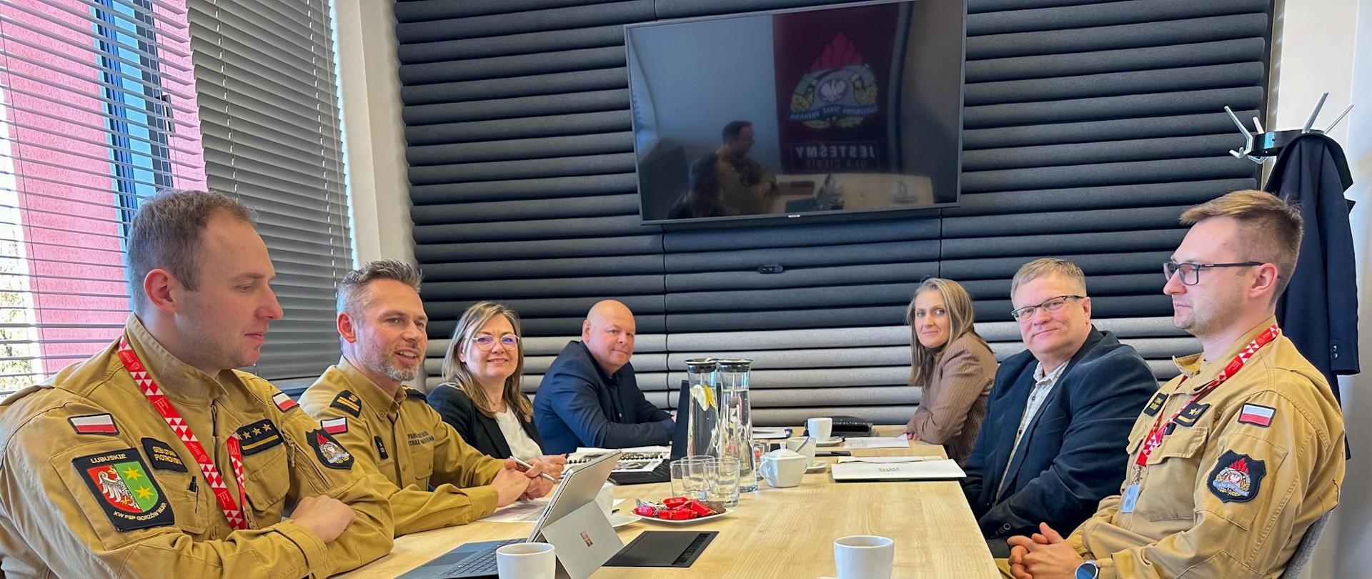 Szerokie ujęcie siedmiu osób zgromadzonych wokół jasnego stołu konferencyjnego podczas spotkania roboczego. Po lewej stronie stołu siedzą trzej strażacy w jasnobrązowych mundurach służbowych z polskimi flagami i insygniami. Pierwszy z nich obsługuje tablet. Za nimi siedzi kobieta i mężczyzna w ubraniach cywilnych. Po prawej stronie stołu siedzą trzy osoby: kobieta w brązowym kostiumie, starszy mężczyzna w sztruksowej marynarce i okularach oraz kolejny strażak w mundurze. Przed uczestnikami stoją filiżanki z kawą, szklanki i dzbanek z wodą. Tło stanowi czarna, rowkowana ściana akustyczna, na której zawieszony jest ekran telewizyjny z logo KW PSP w Gorzowie Wielkopolskim i orłem. Po lewej okno z roletami.