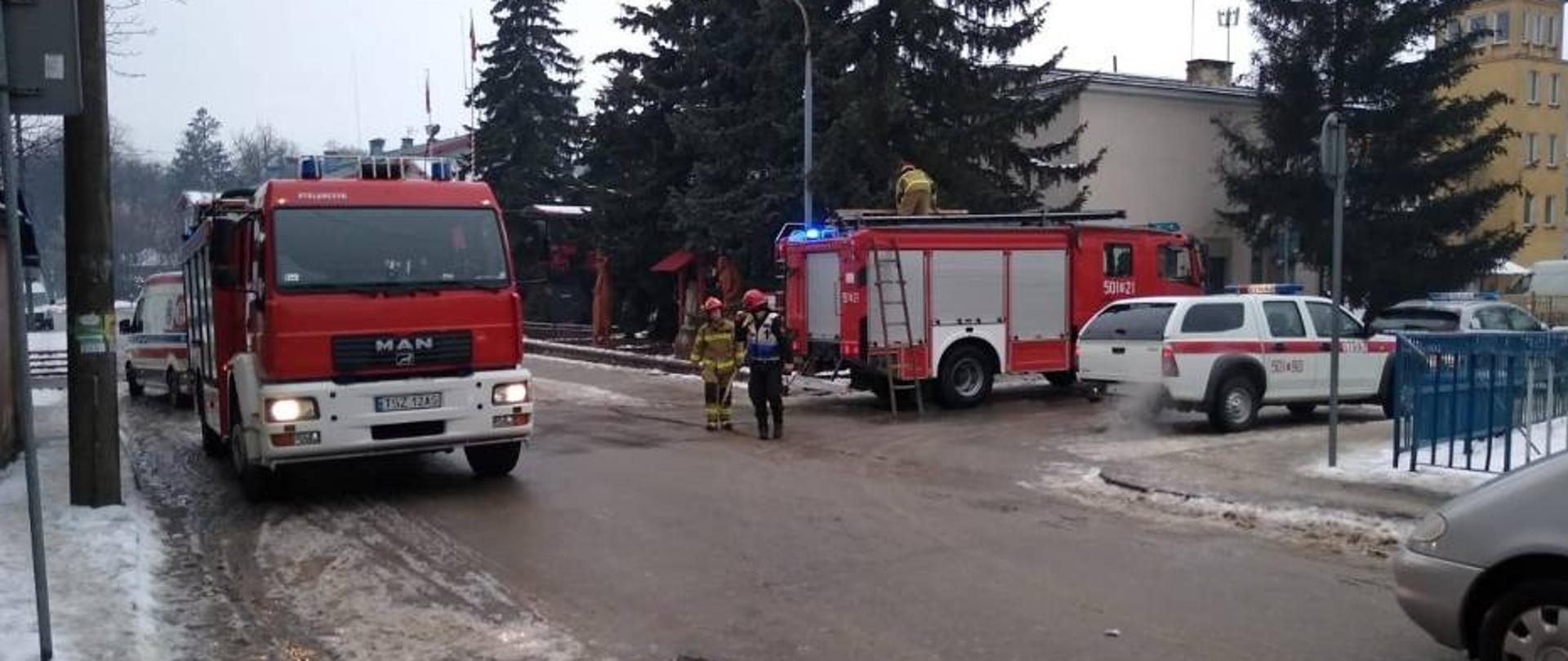 Kierujący działaniami ratowniczymi rozmawia z drugim strażakiem, ubrani są w odzież ochrony osobistej strażaka. Na dachu pojazdu pożarniczego stoi kolejny strażak, wyciąga on sprzęt potrzebny do prowadzenia akcji ratowniczej.
