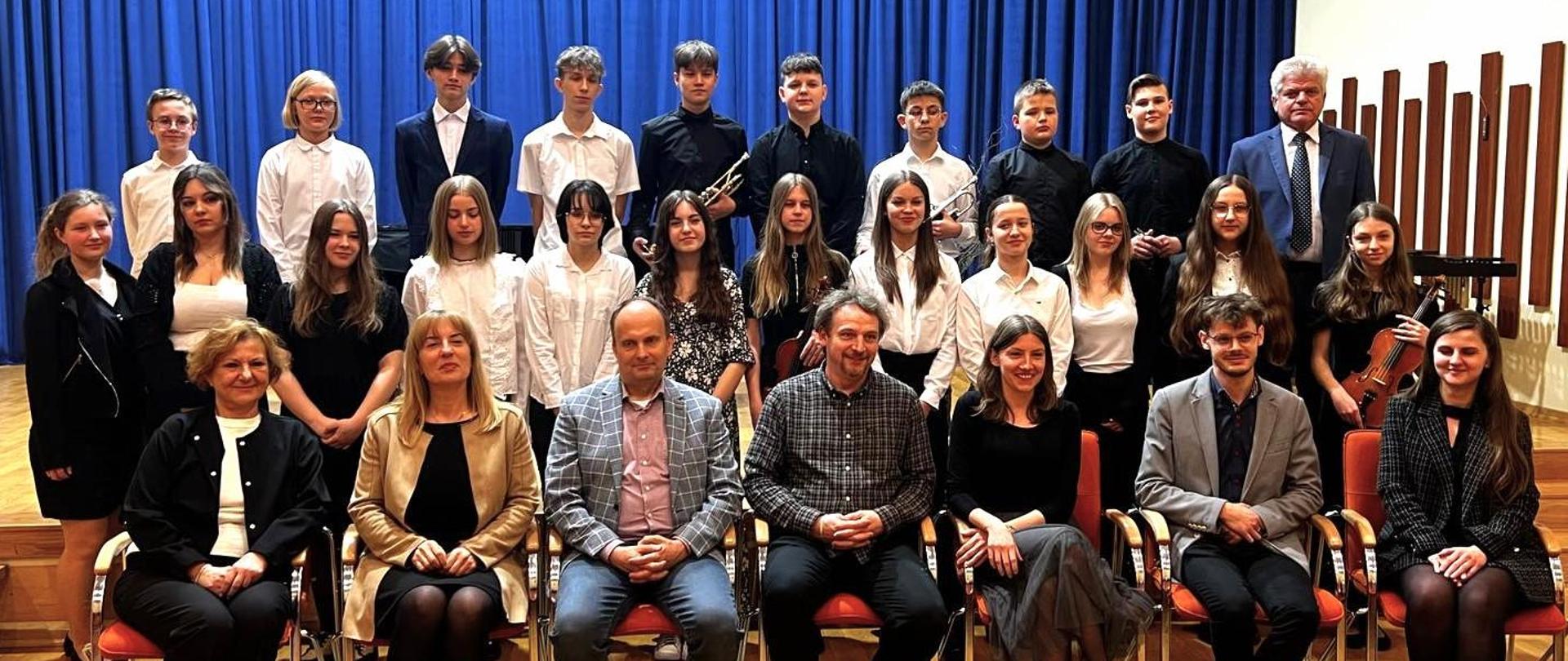 grupa uczniów i nauczycieli na scenie w galowych strojach, w tle niebieska kotara