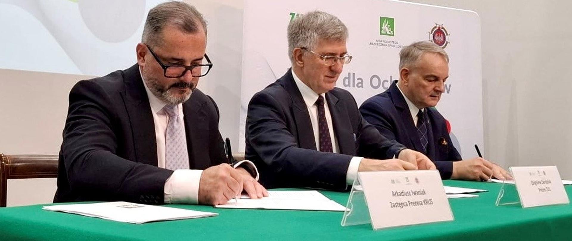 Trzech mężczyzn podpisuje dokumenty przy zielonym stole