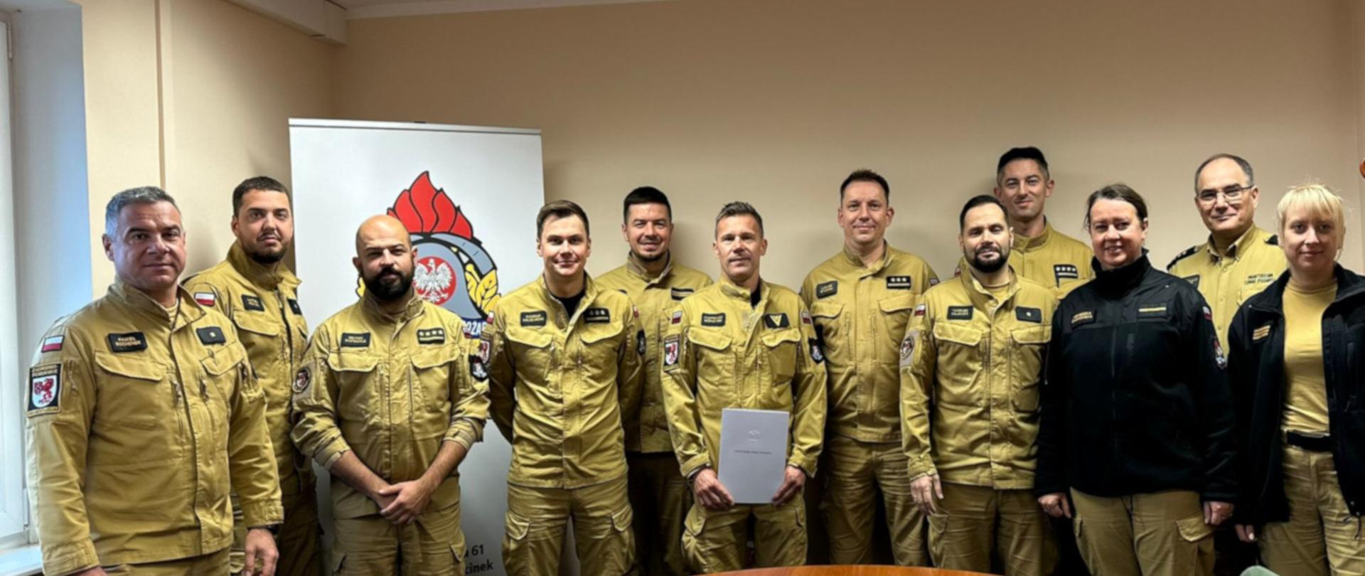 Grupa kilkunastu strażaków i strażaczek w mundurach służbowych stoi razem w pomieszczeniu biurowym, ustawiona w półokręgu za drewnianym stołem. Większość z nich ma na sobie jasnobrązowe mundury, a dwie osoby z prawej strony ubrane są w ciemniejsze, czarne bluzy (polary) służbowe. W centrum grupy znajduje się strażak trzymający jasną teczkę z dokumentem. W tle po lewej stronie stoi roll-up z logo Państwowej Straży Pożarnej. Wszyscy patrzą w stronę aparatu i uśmiechają się lub zachowują neutralny wyraz twarzy, scena rozgrywa się podczas wręczenia nagrody jubileuszowej.