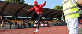 kobieta w czerwonej bluzie i czarnych spodniach z czerwonym hełmie na głowie i prądownicą w ręku pokonuje przeszkodę jaką jest skok przez płotek podczas sztafety kobiet na powiatowych zawodach pożarniczych