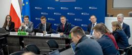 Widok od prawej na salę konferencyjną, stoły ustawione w podkowę. U szczytu drugi od lewej G. Płatek, dalej M. Filipek, M. Jaros. W tle w lewym rogu flagi Polski i UE, obok niebieska ścianka z logotypami MRiT