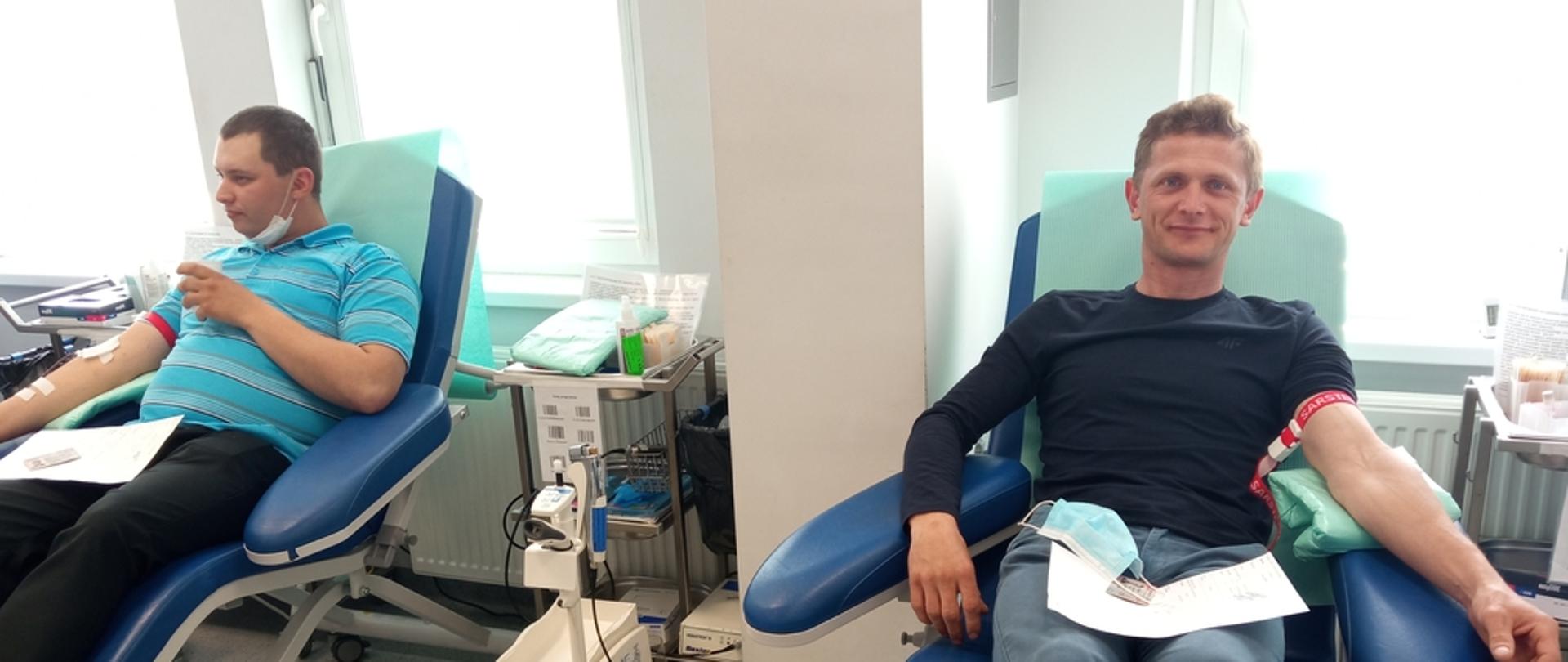 Na zdjęciu dwaj mężczyźni po lewej w niebieskiej koszulce i czarnych spodniach po prawej w czarnej koszulce i dżinsach siedzą na granatowych fotelach oddają krew w pomieszczeniu o białych ścianach i szarej posadzce w Regionalnym Centrum Krwiodawstwa i Krwiolecznictwa w Oddziale Terenowym w Gorlicach.