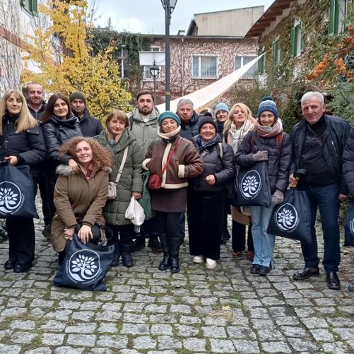 Grupa kobiet i mężczyzn pozuje do zdjęcia z torbami z napisem Machakhela Product, w tle obrośnięte poroślami budynki 