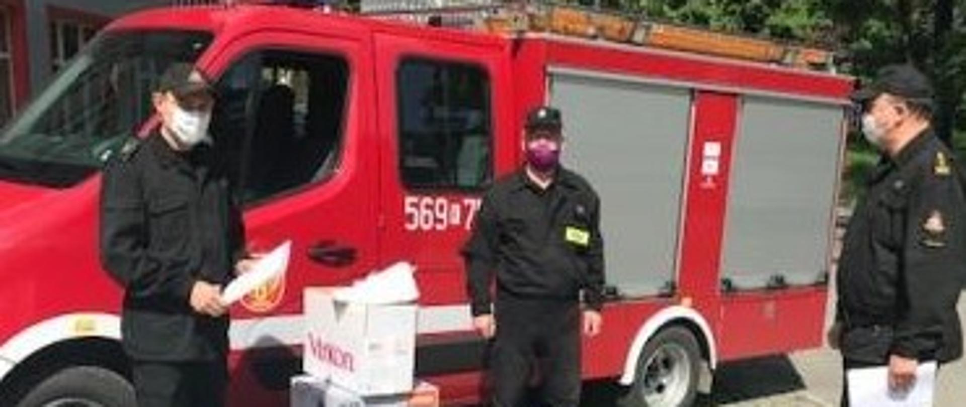 Zastępca Komendanta Powiatowego PSP st. bryg. Marek Rodak wraz z mł. kpt. Bartoszem Kłosowiczem przekazują strażakowi z OSP środki ochrony indywidualnej, płyny wirusobójcze oraz opryskiwacze do działań związanych z COVID-19 na tle lekkiego samochodu ratowniczo - gaśniczego