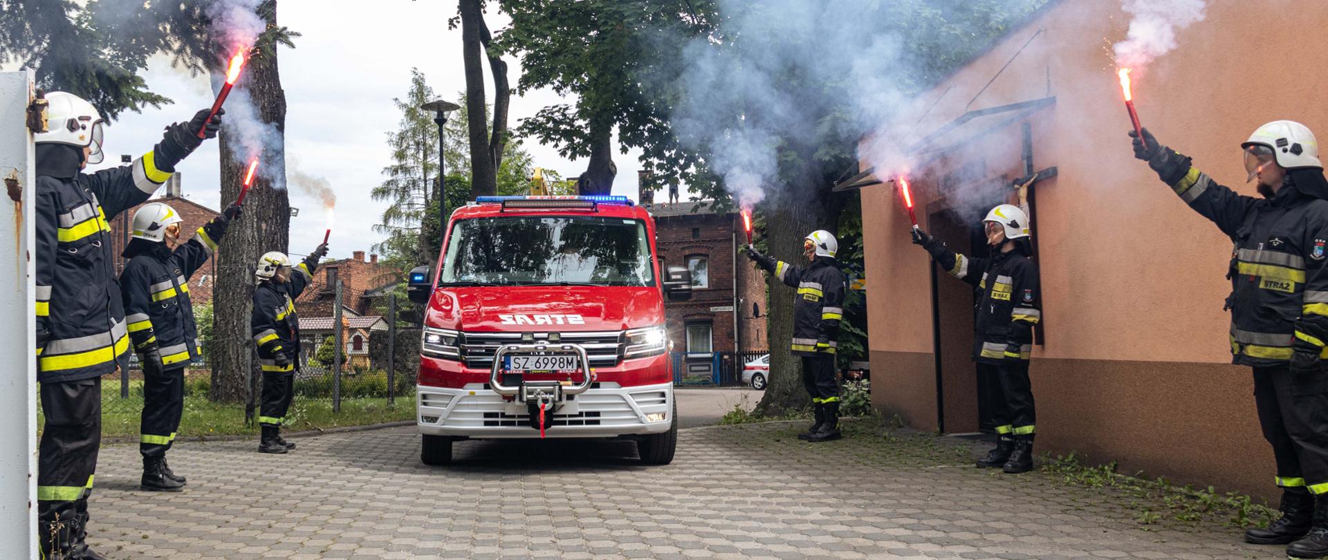 Wjazd nowego lekkiego samochodu ratowniczo-gaśniczego Volkswagen Crafter, z lewej i prawej strony stoi po trzech druhów ubranych w białe hełmy i ubrania specjalne, w ręku trzymają race, z których wydobywa się czerwony płomień i biały dym.