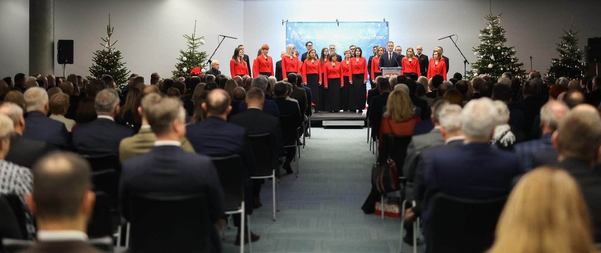 w dwóch rzędach krzeseł siedzi grupa osób, w głębi zdjęcia na scenie stoi grupa kobiet w czerwonych bluzkach i czarnych spódnicach za nimi ciemno ubrani mężczyźni. z prawej strony strony przed nimi stoi mężczyzna przy mównicy