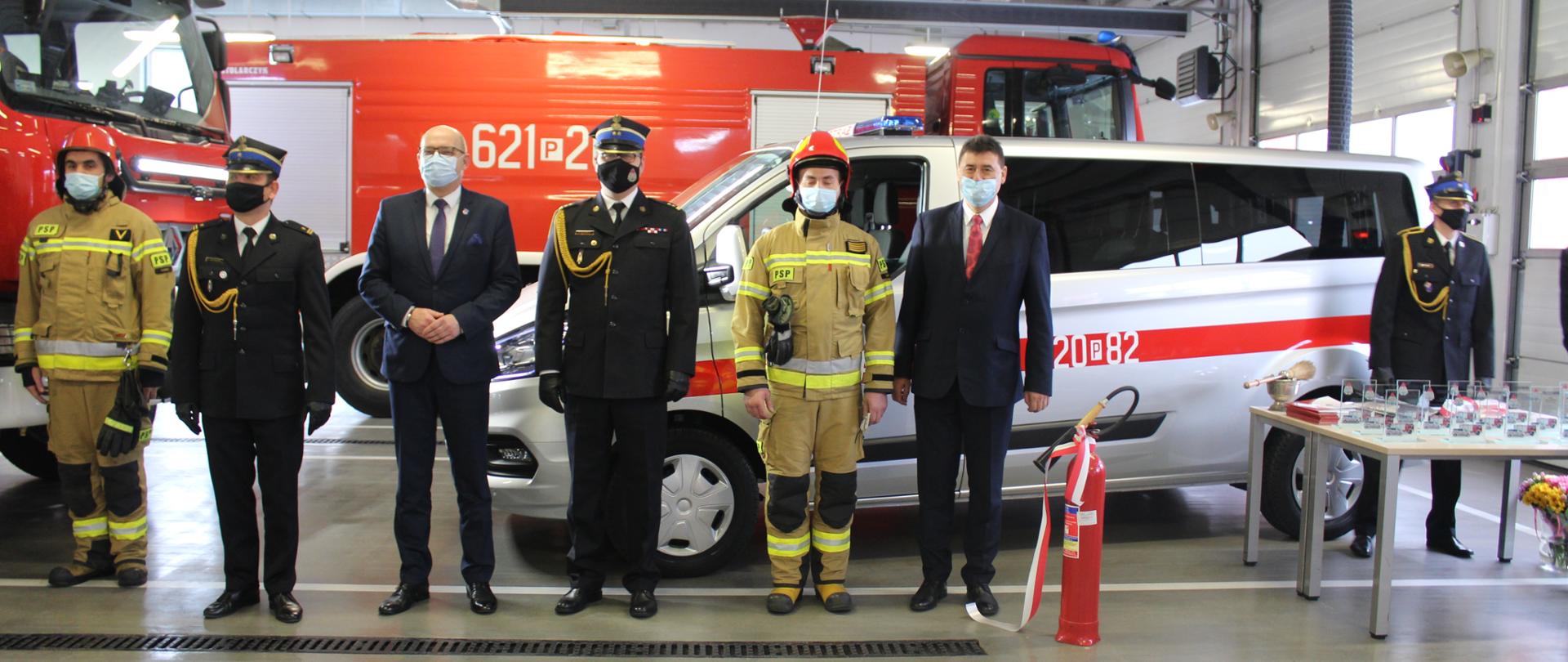 strażacy stoją w szeregu, w tle pojazdy pożarnicze