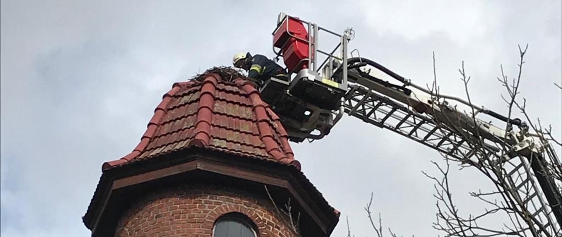 Zdjęcie przedstawia więżę na której znajduję się przechylone bocianie gniazdo oraz drabinę strażacką która znajduje się przy gnieździe celem jego poprawy.