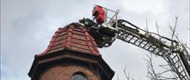 Zdjęcie przedstawia więżę na której znajduję się przechylone bocianie gniazdo oraz drabinę strażacką która znajduje się przy gnieździe celem jego poprawy.