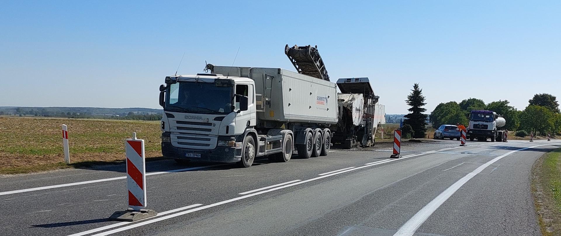 Remont nawierzchni - na jednym pasie ruchu asfaltowej drogi pracuje frezarka. Stoją dwie ciężarówki i samochód osobowy. Pas jezdni zabezpieczony elementami oznakowania biało-czerwonymi.