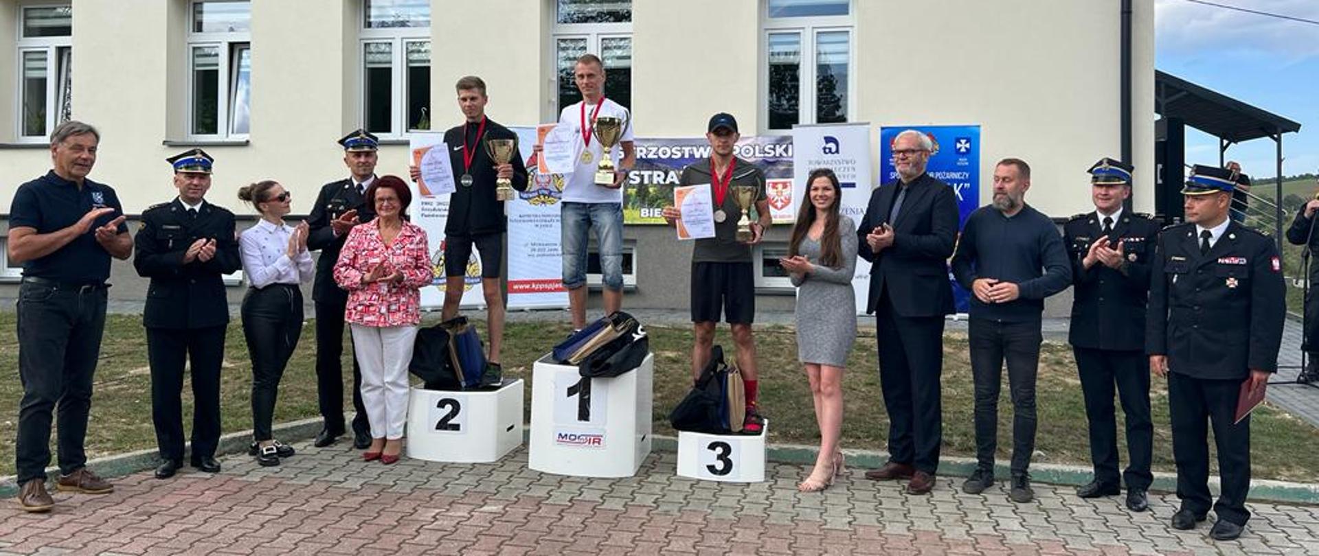 Na podium stoją z pucharami trzej funkcjonariusze, przy podium znajdują się komendanci oraz osoby towarzyszące