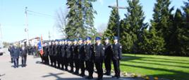 Grupa strażaków ustawionych jeden za drugim stoi na placu wyłożonym kostką. Przed nimi idzie dwóch mężczyzn. Za nimi widać trawnik, krzyż oraz wysokie drzewa. 