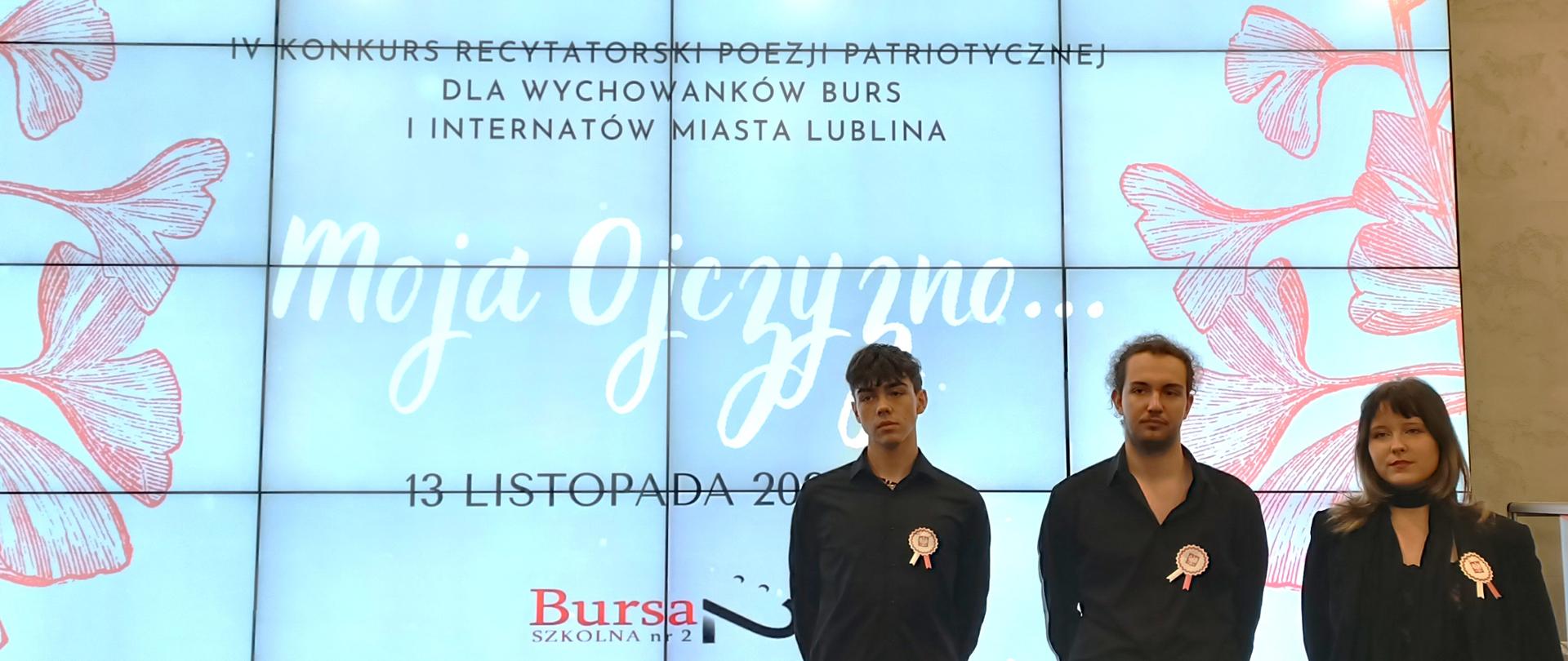 Z prawej strony stoi dwóch chłopców i dziewczyna, wszyscy mają przypięte biało-czerwone kotyliony. Z tyłu jest plansza z napisem "IV KONKURS RECYTATORSKI POEZJI PATRIOTYCZNEJ DLA WYCHOWANKÓW BURS I INTERNATÓW MIASTA LUBLINA MOJA OJCZYZNO...". Powyżej na planszy są logotypy Prezydenta Miasta Lublin z lewej strony i IPN Lublin z prawej strony, a na dole napis 13 listopada 2025 r. i Bursa Szkolna nr 2