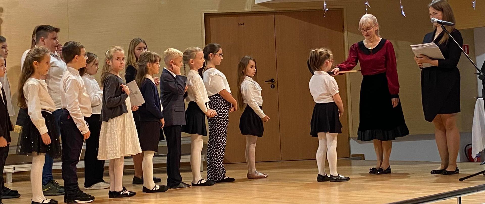 Na scenie uczniowie klas pierwszych są pasowani na pierwszoklasistów przez panią dyrektor wielka nutą.