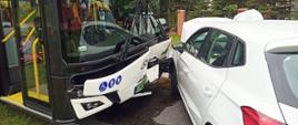 Fotografia przedstawia biały samochód osobowy, który zderzył się z autobusem miejskim oba przody pojazdów są uszkodzone.