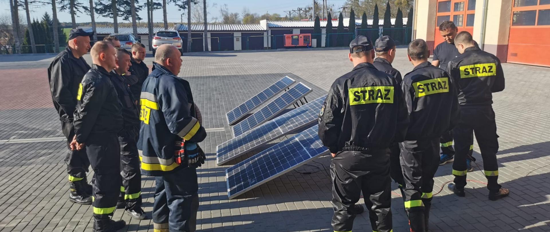 Pora dzienna, na placu KP PSP w Sztumie strażacy ubrani w mundury koszarowe i specjalne stoją wokół położonych na ziemi paneli fotowoltaicznych. Strażak PSP prowadzi instruktarz.