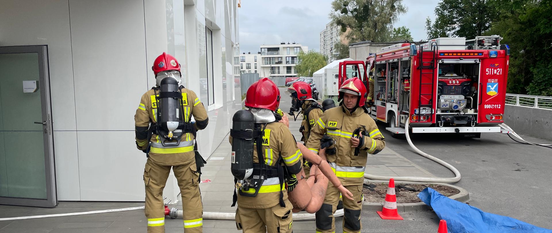 Zdjęcie przedstawia budynek Invicty. Przy budynku stoi samochód strażacki z otwartymi skrytkami, do którego podpięte są węże strażackie. Widać strażaków w ubraniach specjalnych i aparatach powietrznych. Dwóch z nich niesie manekina pozorującego osobę poszkodowaną.