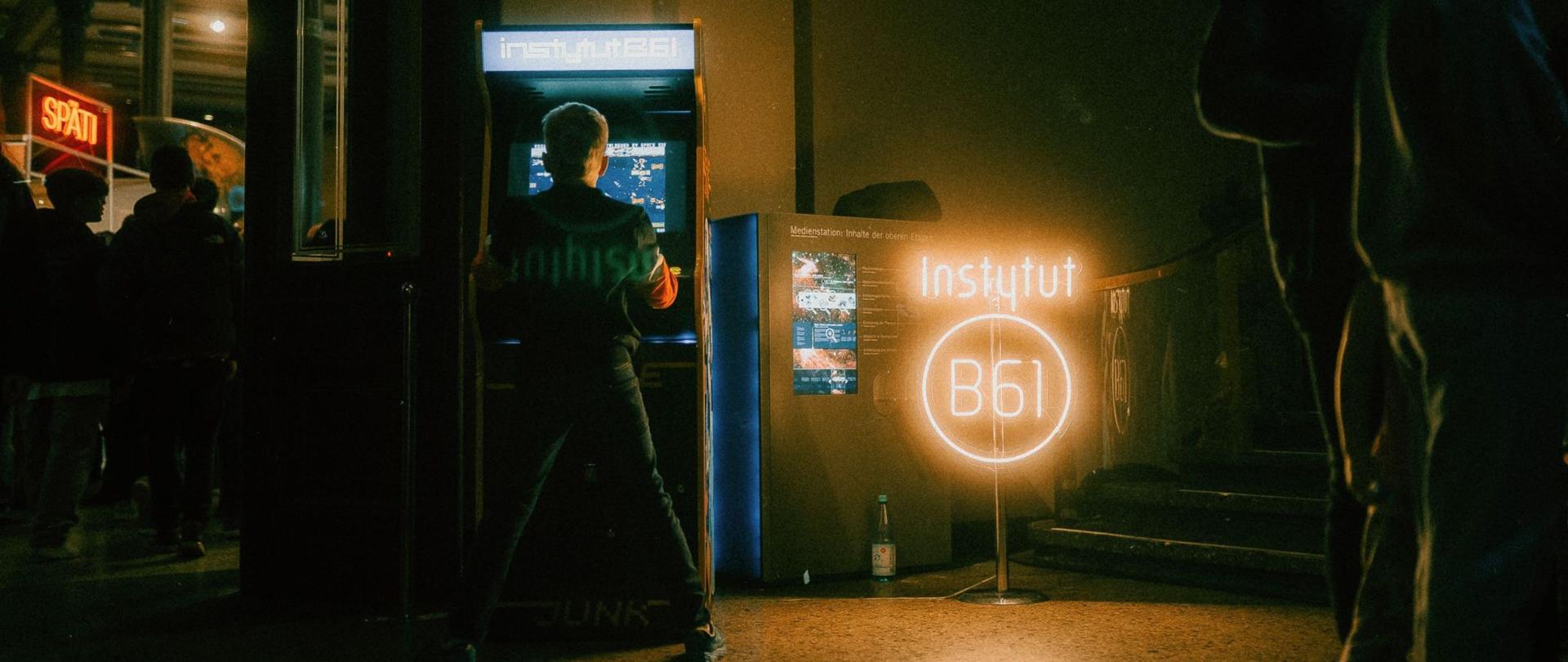 A foto mostra uma criança a jogar numa máquina caça-níqueis. Ao lado, encontra-se um letreiro de néon aceso com as palavras Instytut B61.