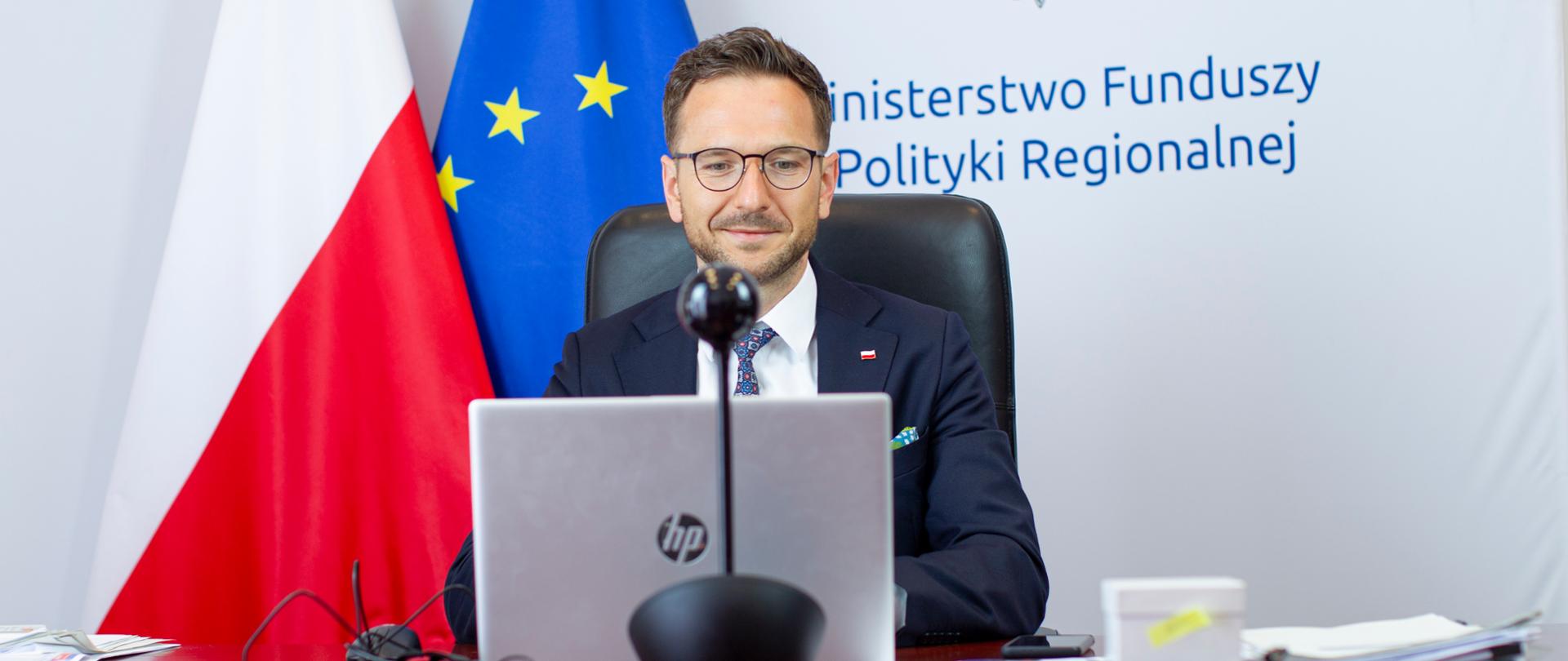 wiceminister Waldemar Buda uczestniczy w spotkaniu online, za nim flagi Polski i Unii Europejskiej