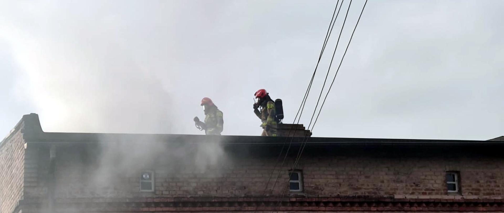 Zdjęcie przedstawia dwóch strażaków na dachu domu jednorodzinnego wyposażonych w sprzęt ochrony układu oddechowego zbliżających się do dymiącego komina.