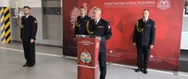 Za mównicą stoi Komendant ubrany w mundur galowy z tyłu stoi trzech strażaków w tle czerwona ścianka 