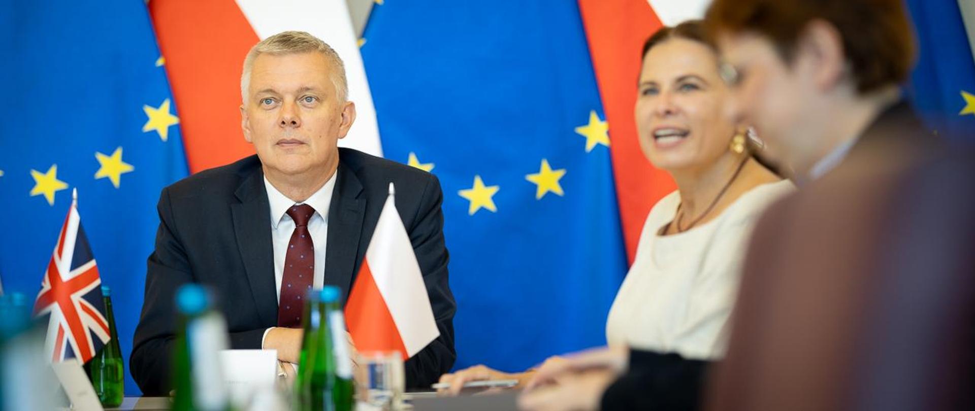 Mężczyzna przy stole, obok flaga Wielkiej Brytanii i Polski. 