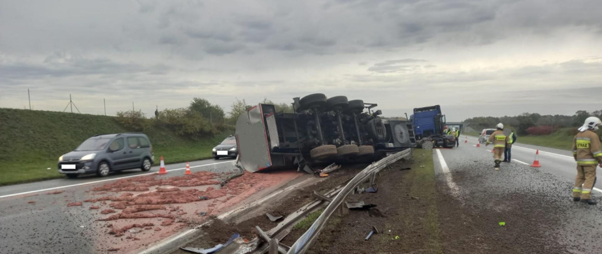 Cysterna leży na drodze, widać rozlaną substancję, po lewej przejeżdża samochód osobowy, po prawej widać dwóch strażaków