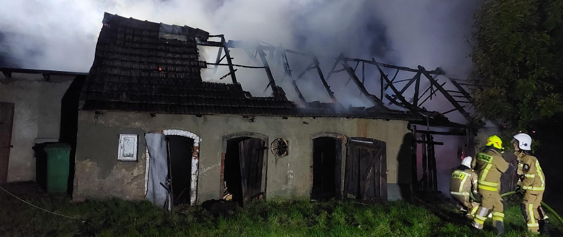 Jest pora nocna, przed budynkiem trzech strażaków rozwija linię gaśniczą. Na zdjęciu widać budynek gospodarczo-inwentarski nad którym unosi się zadymienie. Większa część dachu jest już spalona