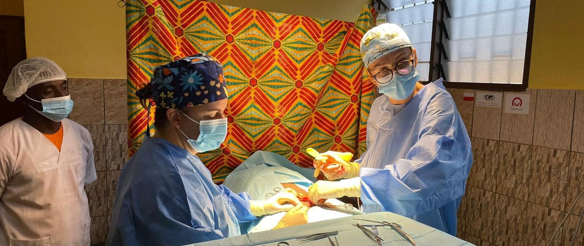 Kobieta i mężczyzna w fartuchach, czapkach i maseczkach stoją przy stole i trzymają narzędzia chirurgiczne. Trzeci mężczyzna ubrany tak samo przygląda się. 