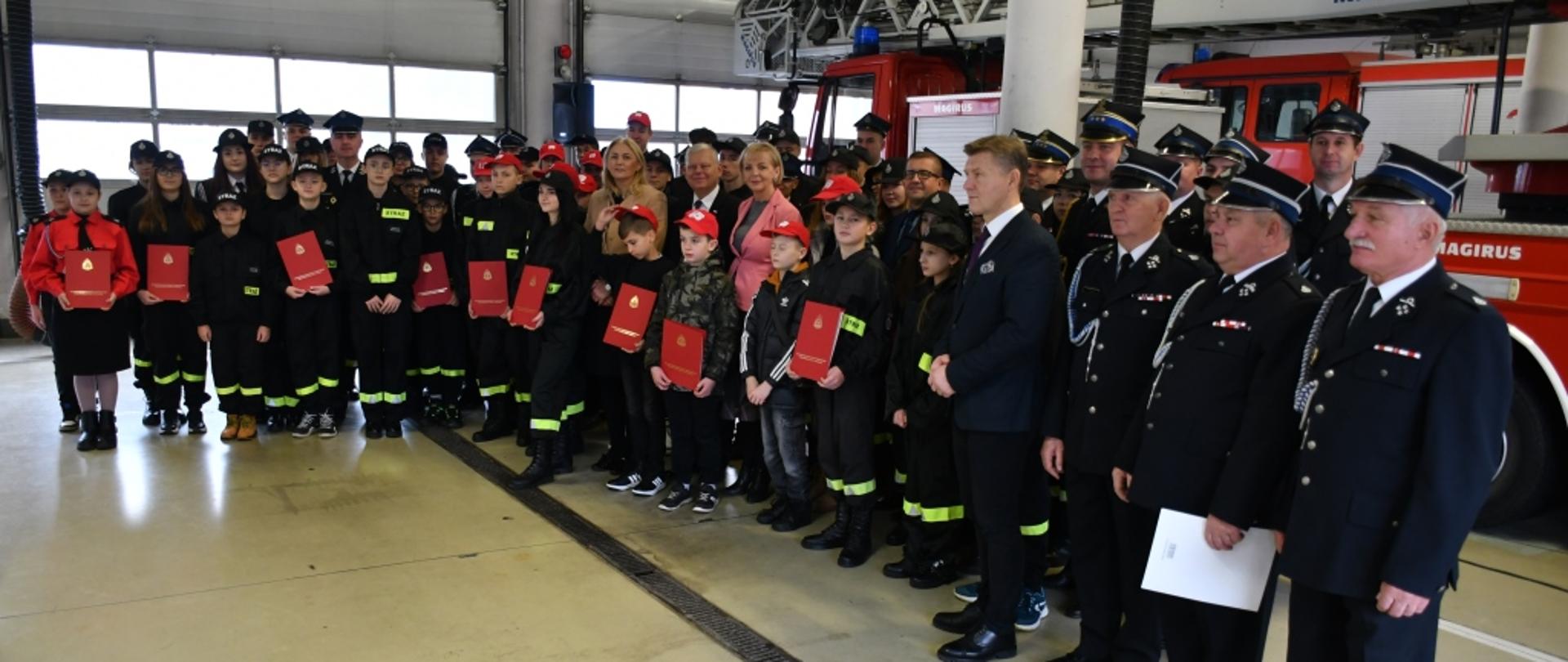 Pamiątkowe zdjęcie upamiętniające uroczystość wręczenia promes dla Młodzieżowych Drużyn Pożarniczych. Na zdjęciu ustawieni są obok siebie są członkowie drużyn, funkcjonariusze Państwowej i Ochotniczej Straży Pożarnej, posłowie, samorządowcy. Zdjęci wykonane na terenie jednostki ratowniczo – gaśniczej Państwowej Straży Pożarnej numer 1 w Radomiu. Za zebranymi w tle znajduje się pojazd pożarniczy drabina mechaniczna. 