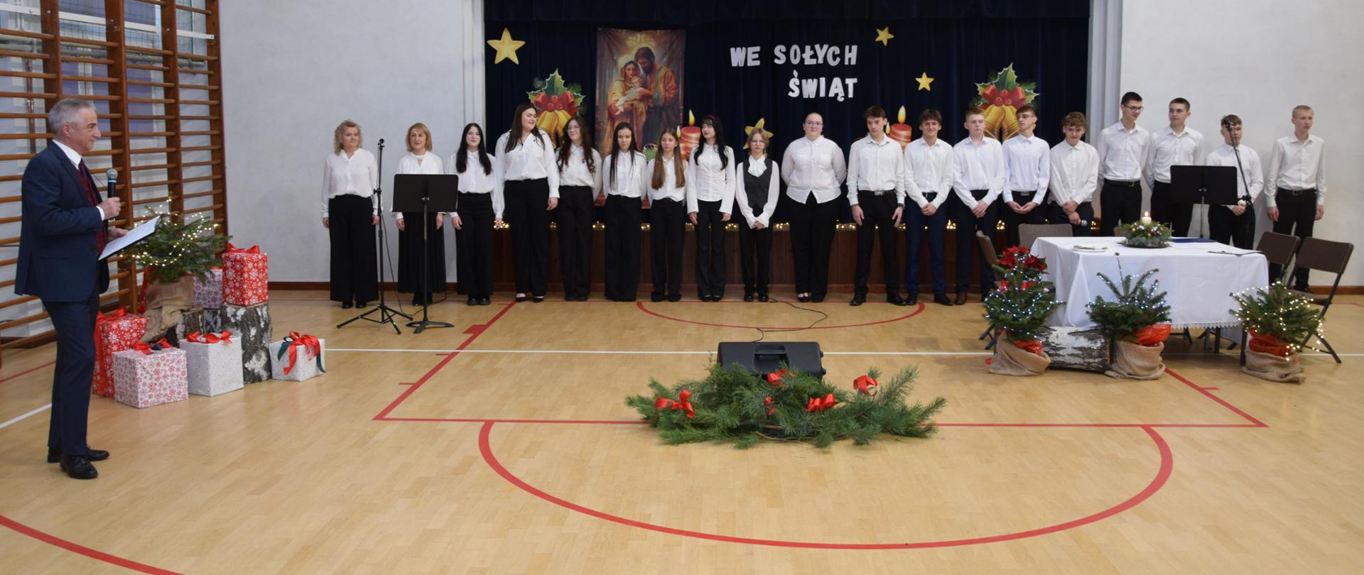 Artyści wraz z opiekunkami stoją w rzędzie, przed nimi dyrektor z mikrofonem, dekoracje świąteczne 