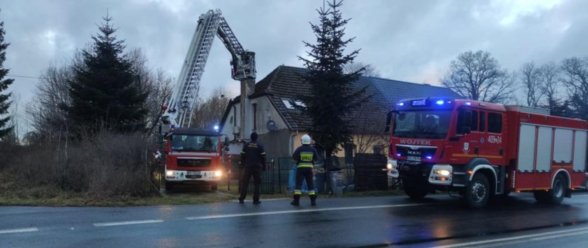 Zdjęcie przedstawia działania gaśnicze. Widać na nim stojący przy budynku podnośnik strażacki oraz drugi wóz gaśniczy. Przed budynkiem stoi strażak w białym hełmie oraz strażnik miejski. Auta mają włączone niebieskie światła ostrzegawcze. Trwa akcja gaśnicza